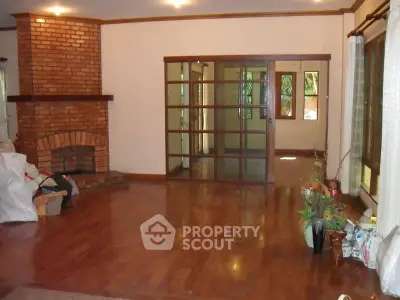 Spacious living room with wooden floors and brick fireplace, featuring large glass doors leading to a sunlit area.