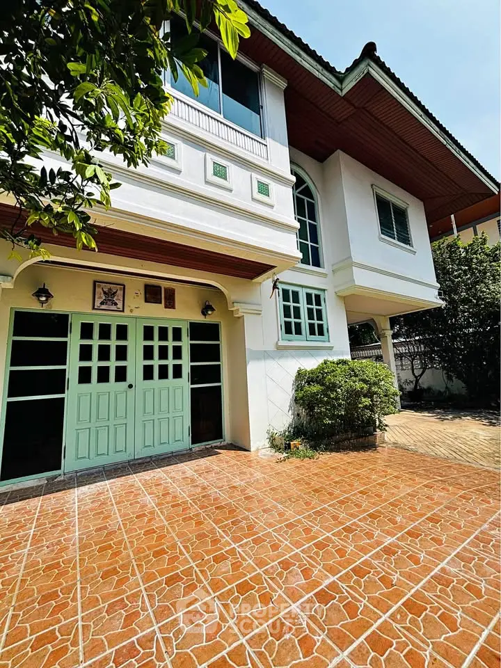 Charming two-story house with green accents and spacious tiled driveway, surrounded by lush greenery.