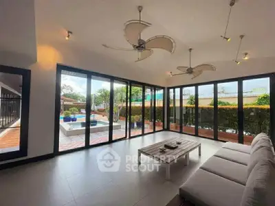 Spacious living room with glass walls overlooking a pool and garden, featuring modern ceiling fans.