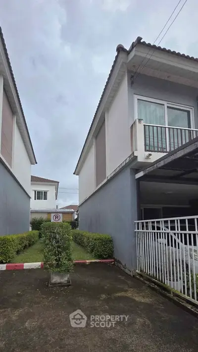 Modern two-story house with a small balcony and driveway, surrounded by greenery.