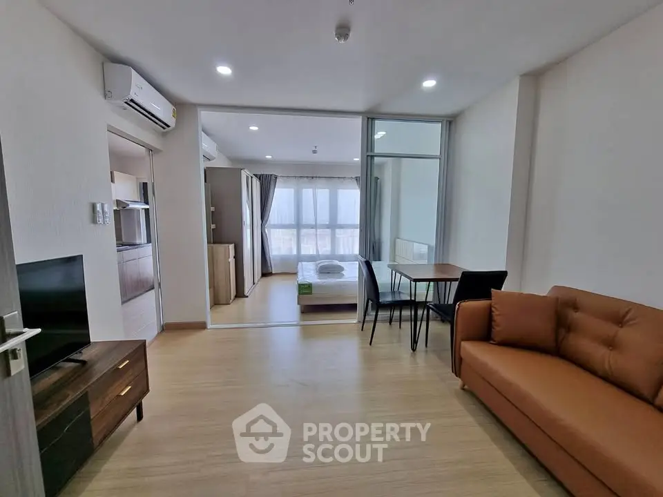 Modern open-plan living room with cozy sofa and dining area, leading to a bright bedroom with large windows.