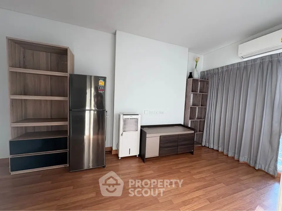 Spacious modern living room with wooden flooring and sleek furniture, featuring a stainless steel fridge and air conditioning.