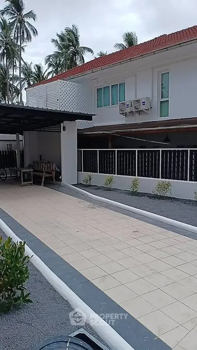 Charming white house with red roof, spacious driveway, and lush palm trees in tropical setting.