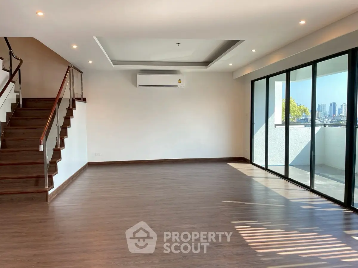 Spacious modern living room with large windows and city view, featuring wooden flooring and elegant staircase.