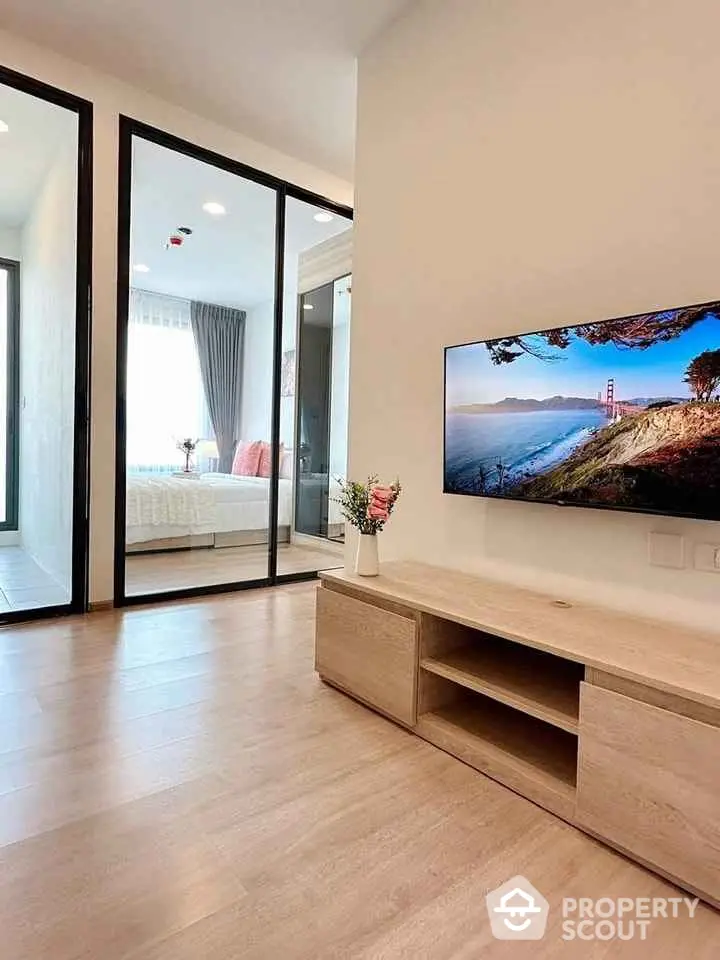 Modern living room with wooden flooring and TV, adjacent to a bright bedroom with glass partition.