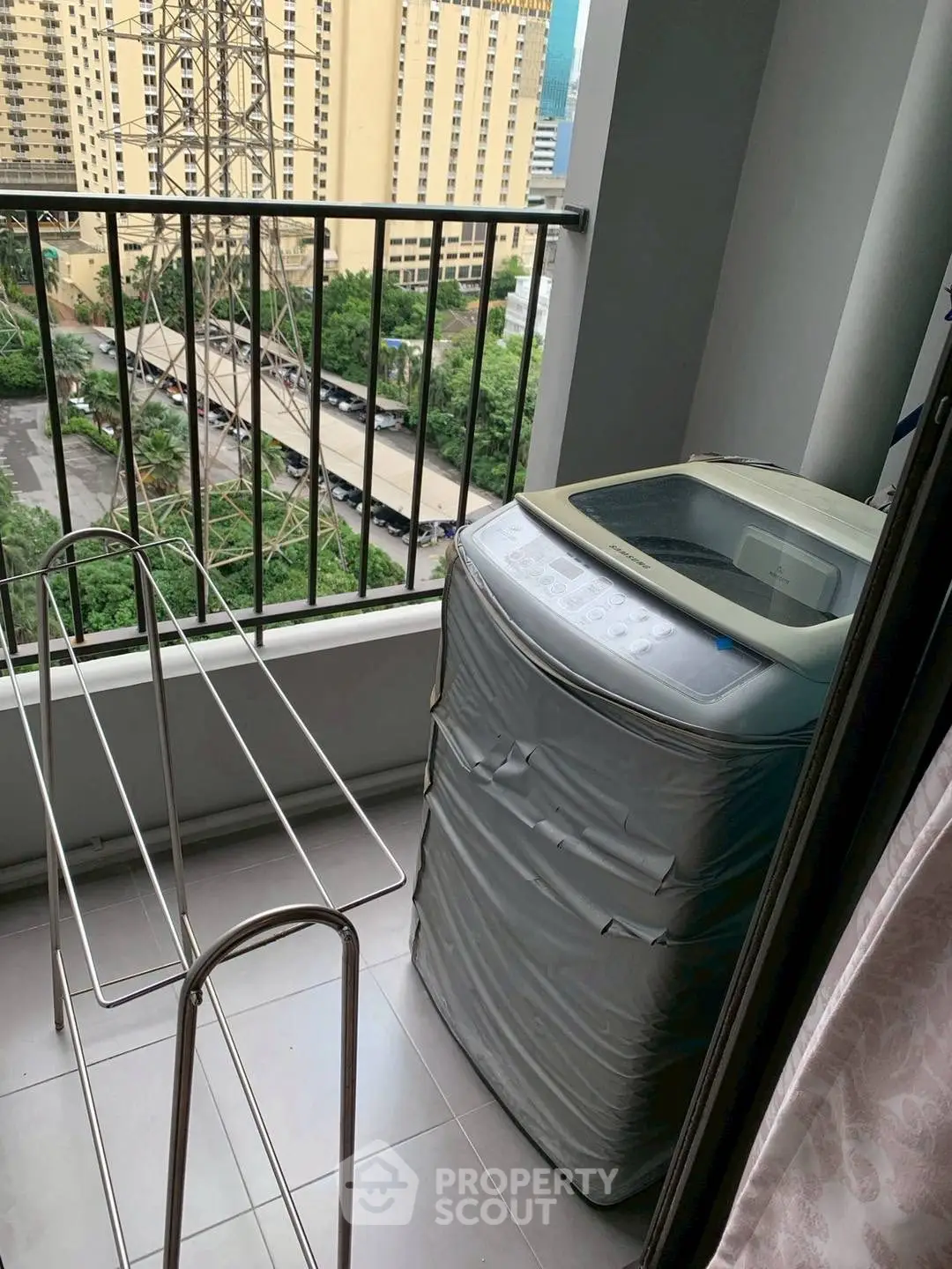 Compact balcony with washing machine and drying rack, city view in background.