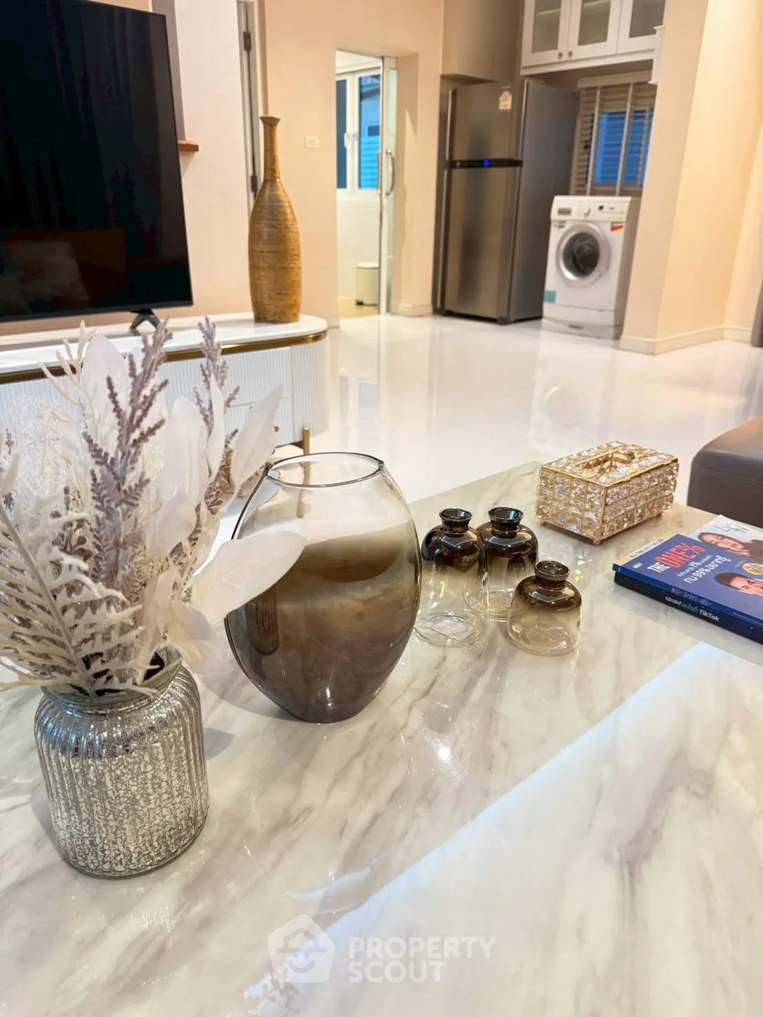 Modern living room with elegant decor, featuring a marble table, stylish vases, and a view of the kitchen with appliances.