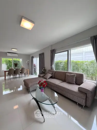 Spacious modern living room with elegant sofa and dining area, featuring large windows and natural light.