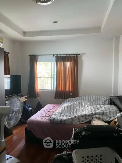 Cozy bedroom with natural light and wooden flooring, featuring a TV and fan for comfort.