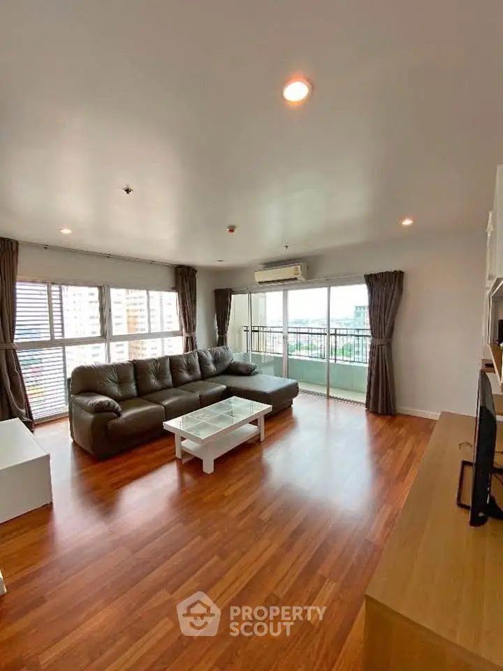 Spacious living room with large windows and city view, featuring modern furniture and wooden flooring.