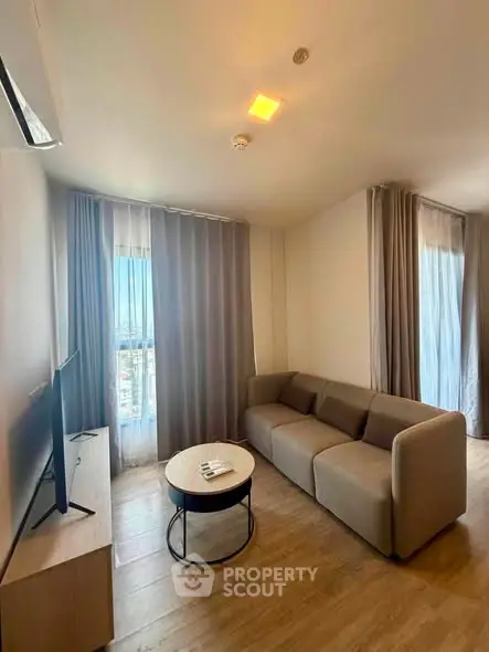 Modern living room with cozy sofa and flat-screen TV, featuring large windows and elegant curtains.
