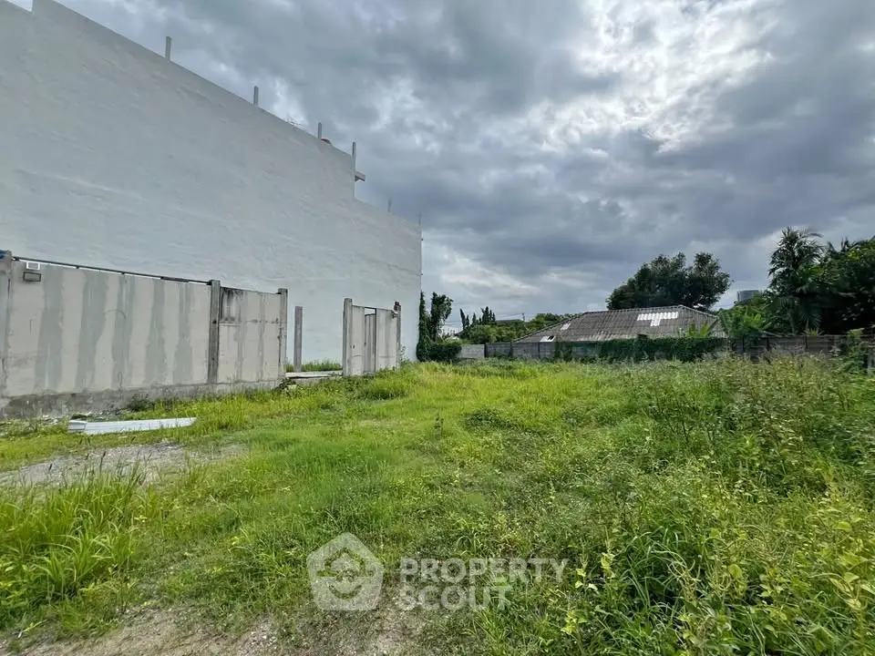 Vacant land with lush greenery and adjacent building under cloudy sky, ideal for development.