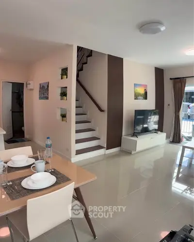 Modern living room with staircase, dining area, and large TV, featuring sleek design and natural light.