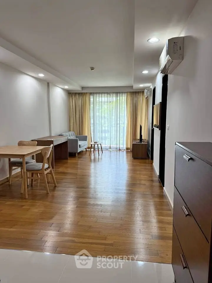Spacious living room with wooden flooring and modern furniture, featuring a large window with natural light.