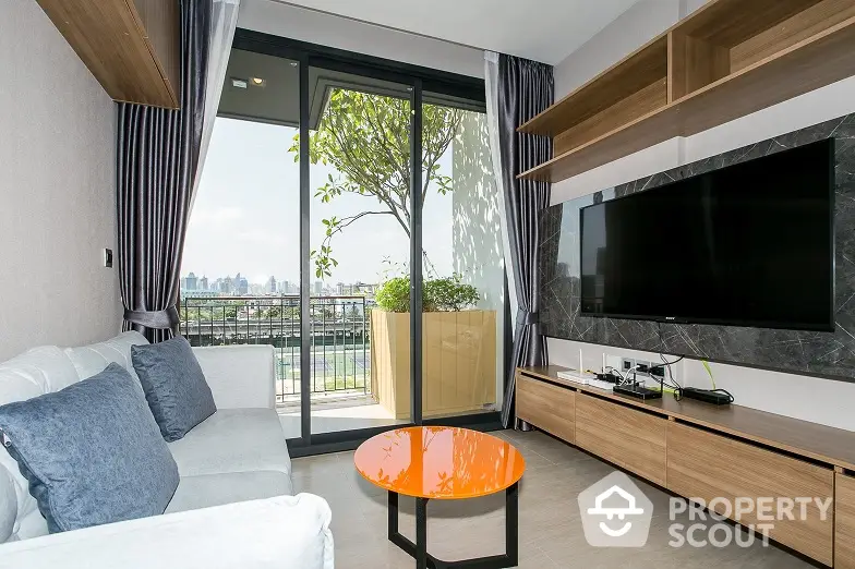 Modern living room with sleek furnishings, large flat-screen TV, and floor-to-ceiling windows leading to a sunny balcony with an urban view.