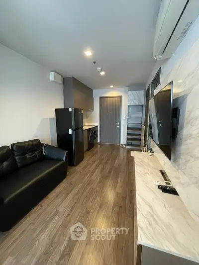 Modern living room with sleek design, featuring a black leather sofa, wall-mounted TV, and compact kitchen area.