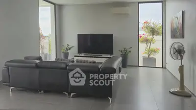 Spacious modern living room with sleek black leather sofa and large TV, featuring floor-to-ceiling windows and lush greenery.