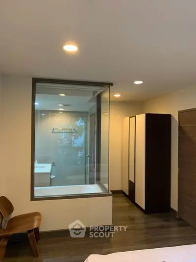 Modern bedroom with glass wall view into bathroom featuring bathtub and sleek design.