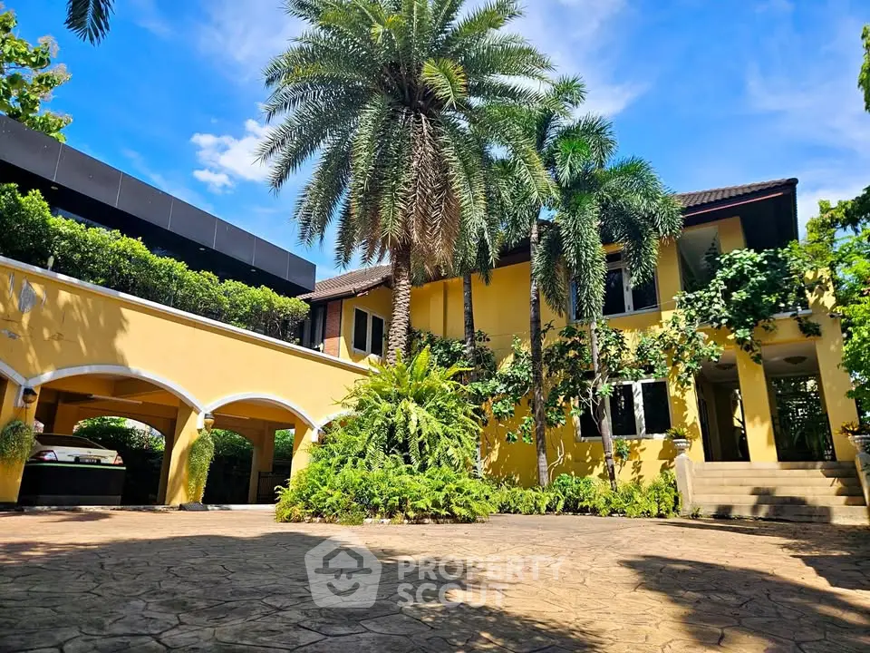 Stunning yellow villa with lush greenery and palm trees, featuring a grand entrance and spacious driveway.