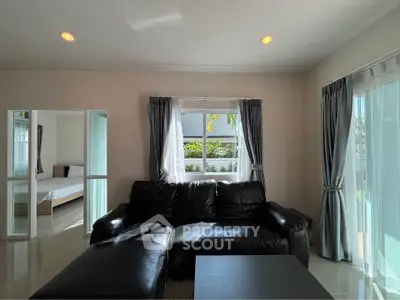 Spacious living room with black leather sofa and natural light, adjacent to a cozy bedroom.