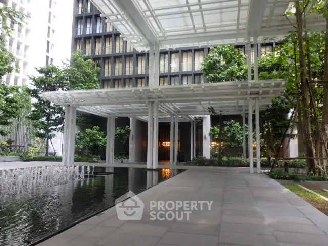 Modern building entrance with sleek design and water feature, surrounded by lush greenery.