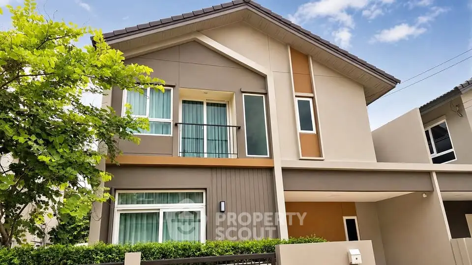 Modern two-story house with large windows and a small balcony, surrounded by greenery and a clear blue sky.
