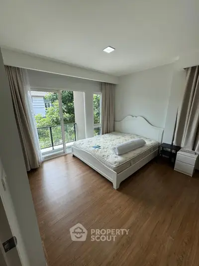 Spacious bedroom with large windows and wooden flooring, offering a serene view of greenery.