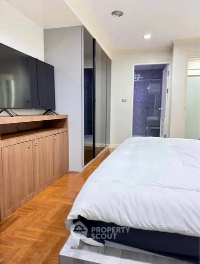 Spacious bedroom with modern decor and large TV, featuring sleek wooden flooring and ample storage space.