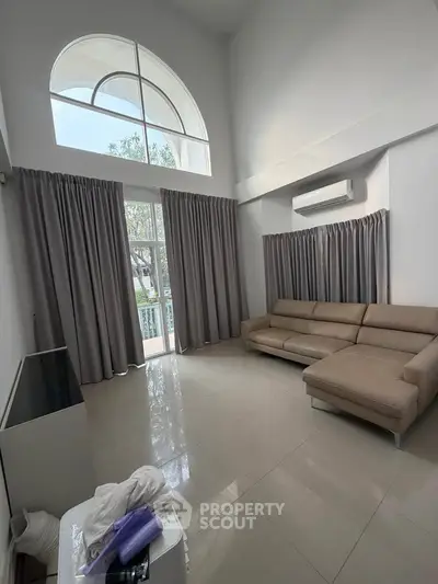 Spacious living room with high ceiling and large windows, featuring modern sofa and air conditioning.