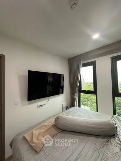 Modern bedroom with large windows and wall-mounted TV, offering a serene view of greenery.