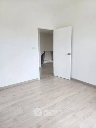 Spacious empty room with light wood flooring and open door leading to hallway.