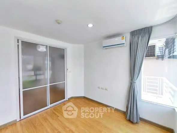 Bright empty room with wooden flooring and large window, featuring air conditioning and sliding door.