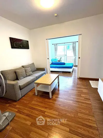 Spacious living room with wooden flooring and cozy sofa leading to a bright bedroom with large windows.