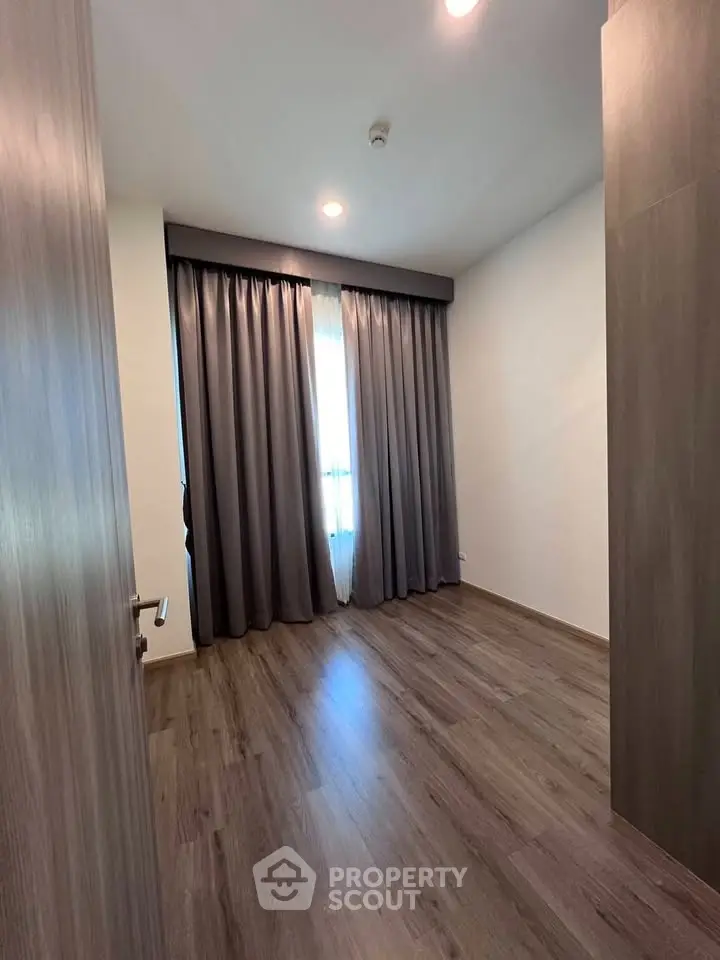 Spacious empty bedroom with wooden flooring and large window with curtains.