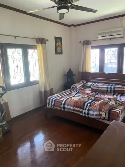 Cozy bedroom with wooden flooring and large windows, featuring a ceiling fan and air conditioning.