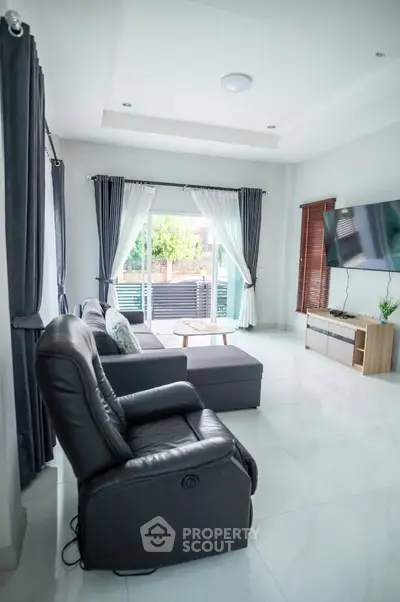 Spacious modern living room with plush leather sofa, sleek entertainment unit, and abundant natural light streaming through large glass doors leading to a cozy outdoor seating area.
