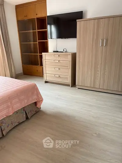 Spacious bedroom with modern furniture and wooden flooring, featuring a wall-mounted TV and ample storage space.