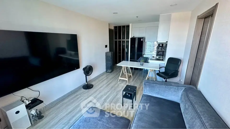 Modern living room with sleek furniture and large TV, open layout to kitchen area.