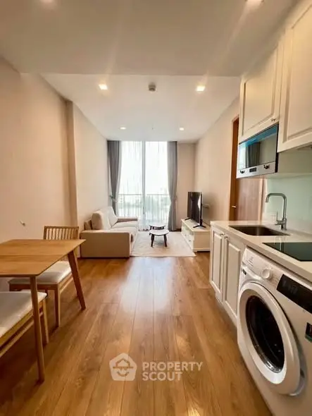 Modern living room with open kitchen and washing machine, featuring wooden flooring and natural light.