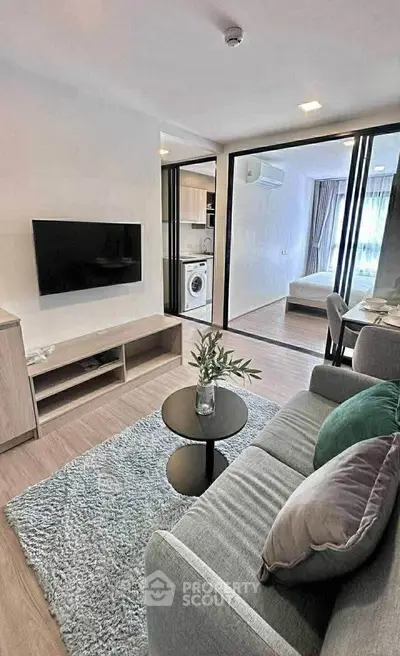Modern living room with open layout, featuring a cozy sofa, flat-screen TV, and view into kitchen and bedroom.
