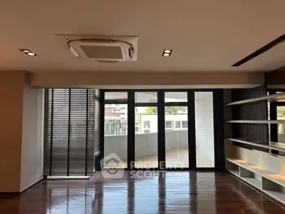 Spacious living room with large windows and balcony access, featuring modern shelving and sleek wooden flooring.