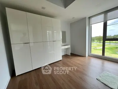 Spacious and modern living room with large windows offering a serene golf course view, sleek white storage cabinets, and rich wooden flooring.