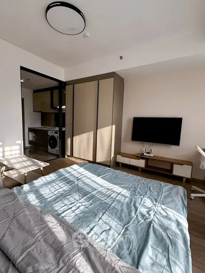 Modern bedroom with sleek design, featuring a cozy bed, wall-mounted TV, and a compact kitchen area with washing machine.