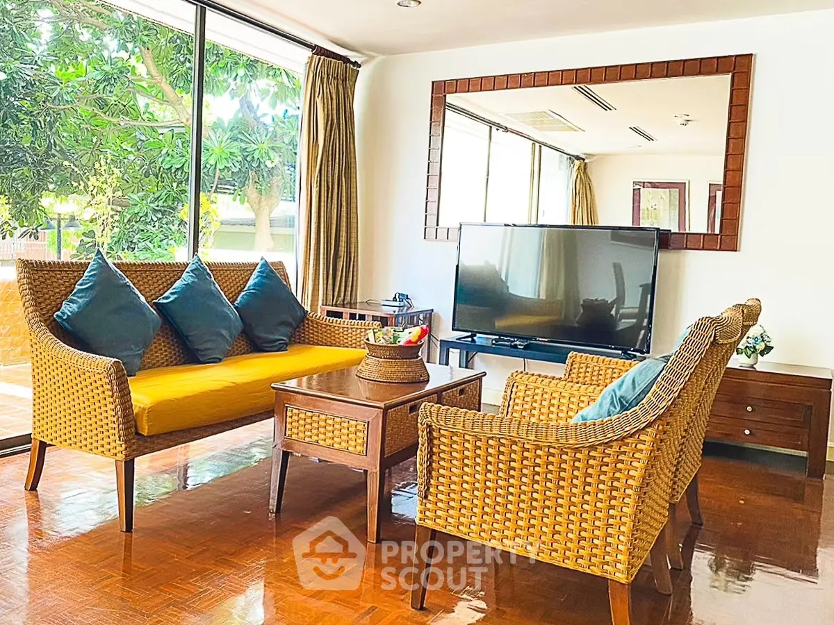 Charming living room with wicker furniture and large windows offering lush garden views.