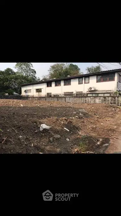 Spacious land plot with potential for development, featuring a modest building and lush greenery in the background.