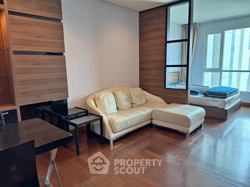 Modern open-layout living room with cozy sofa and bedroom view, featuring wooden flooring and large windows.