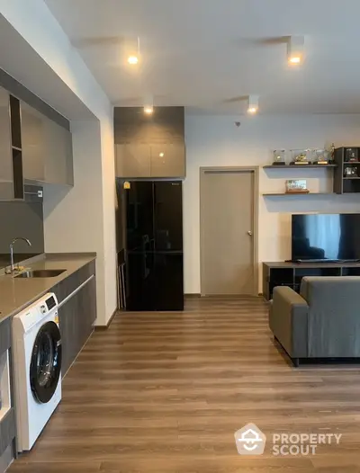 Modern living room with sleek kitchen and washing machine, featuring contemporary design and ample lighting.