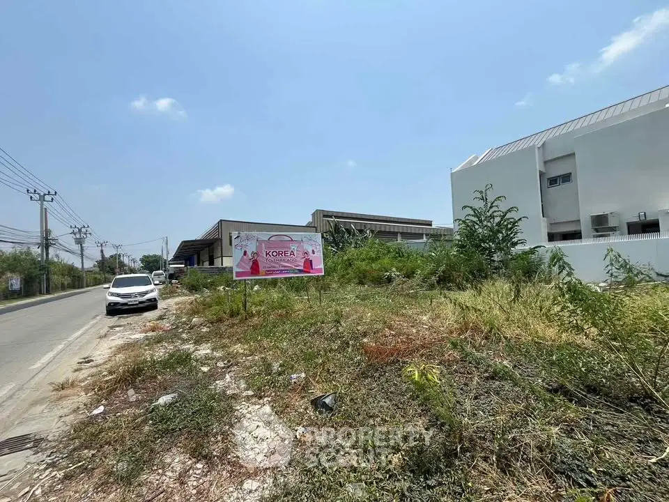 Vacant land plot with road access and nearby buildings under clear blue sky.