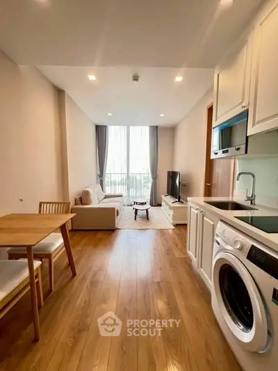 Modern living room with open kitchen and washing machine, featuring wooden flooring and natural light.