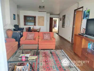 Spacious living room with polished hardwood floors, vibrant orange sofas, and a large glass coffee table, leading to a cozy hallway.